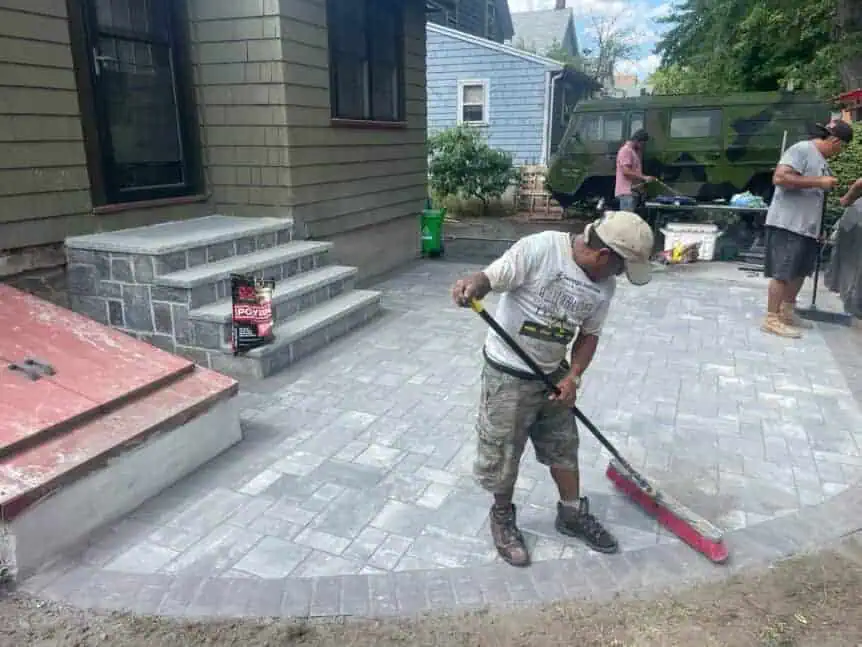 Professional landscapers installing interlocking pavers in a residential driveway or patio in Cambridge. Workers are carefully placing rectangular and square pavers on a leveled sand and gravel base, ensuring even spacing and a smooth, level surface. Construction tools, including rubber mallets, wheelbarrows, and measuring devices, are visible nearby. The background shows landscaped garden beds and a residential home, highlighting precise craftsmanship in outdoor hardscaping.