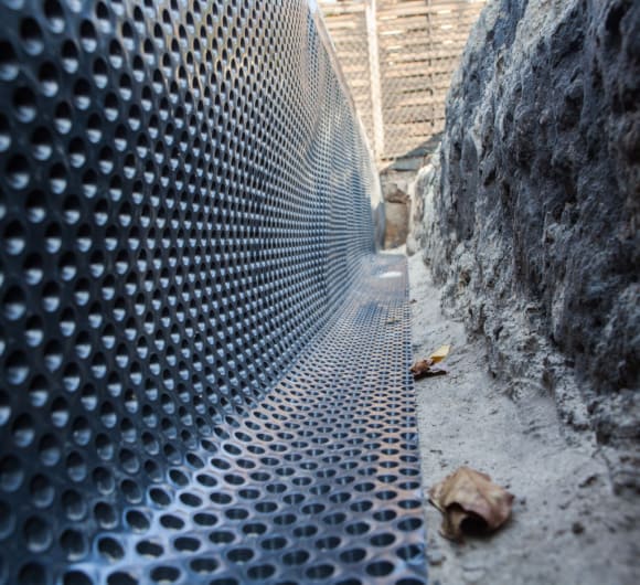 Construction worker applying a waterproofing membrane to the interior walls of a basement in a Cambridge residential property. The worker is using a roller to coat concrete walls with a protective barrier against water infiltration. The basement floor is prepared with drainage channels, and construction tools and materials are arranged nearby. The scene emphasizes professional techniques to prevent basement flooding and moisture damage.