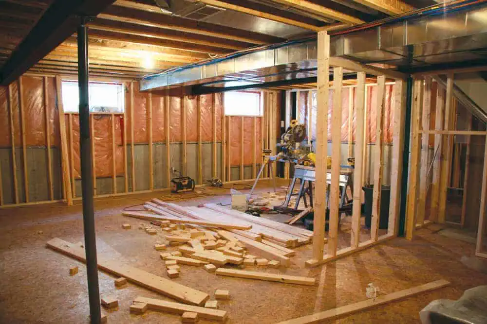A basement under construction with exposed wooden beams and insulation on the walls. A circular saw and other tools are on a table, and scattered wood pieces are on the floor. There is partial framing for future walls.