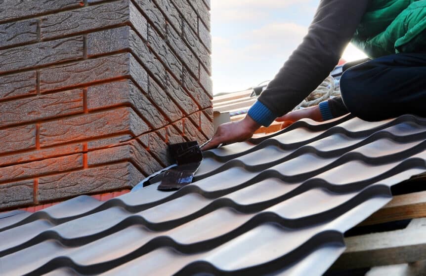 A roofer or builder attaching a metal sheet to an unfinished chimney on a house roof. The worker is using tools to secure the metal flashing, which will protect the chimney from water infiltration once the roof and chimney are completed. The roof surface and partially constructed chimney are visible, highlighting professional roofing and waterproofing work in progress.