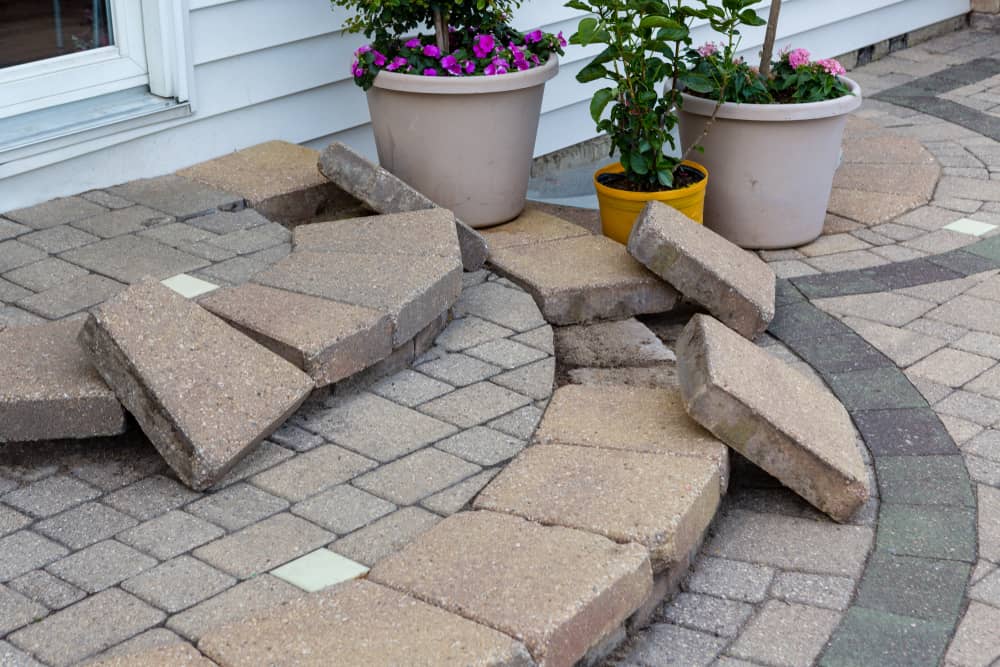 A pathway made of stone tiles is damaged and uneven, with several stones missing or displaced. Three potted plants with vibrant flowers are placed near the path, which could benefit from professional paver installations in Boston, next to a light-colored building with white siding.