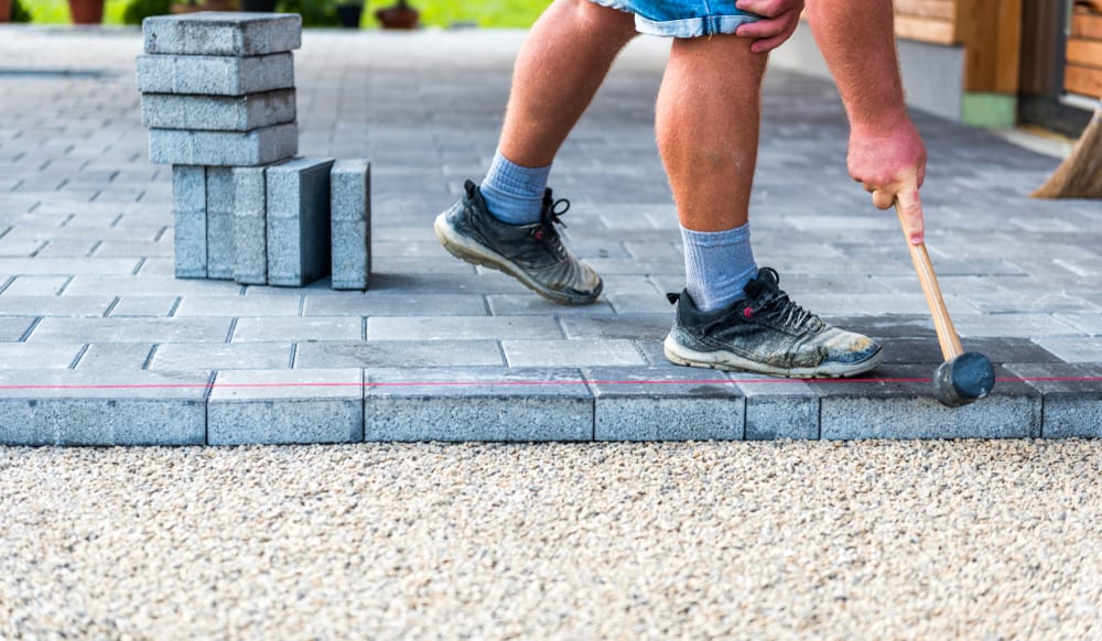 A person wearing sneakers, shorts, gray socks, and a blue shirt uses a rubber mallet to lay gray paving stones on a patio. A stack of additional stones is nearby, and gravel is visible around the paving area, perfect for showcasing expert paver installations in Boston.