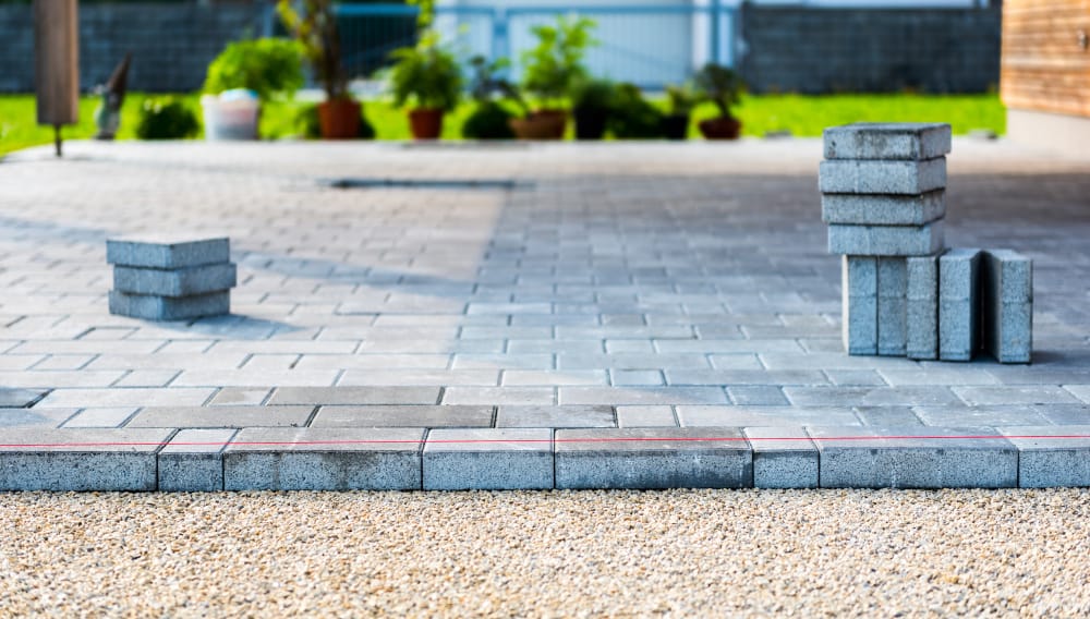 A freshly laid stone patio with stacks of pavers on it, surrounded by a gravel area in the foreground. In the background, there are potted plants and a wooden structure. These Paver Installations Boston are arranged in a neat, orderly manner, enhancing the serene environment.