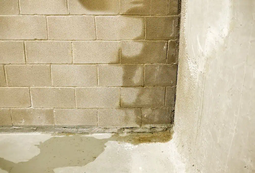 A concrete wall with visible water damage near the bottom corner. The brick surface shows darker patches indicating dampness, while the adjacent plain concrete surface appears less affected. The floor also shows a damp area extending from the corner, suggesting a potential need for Foundation Repair Boston.