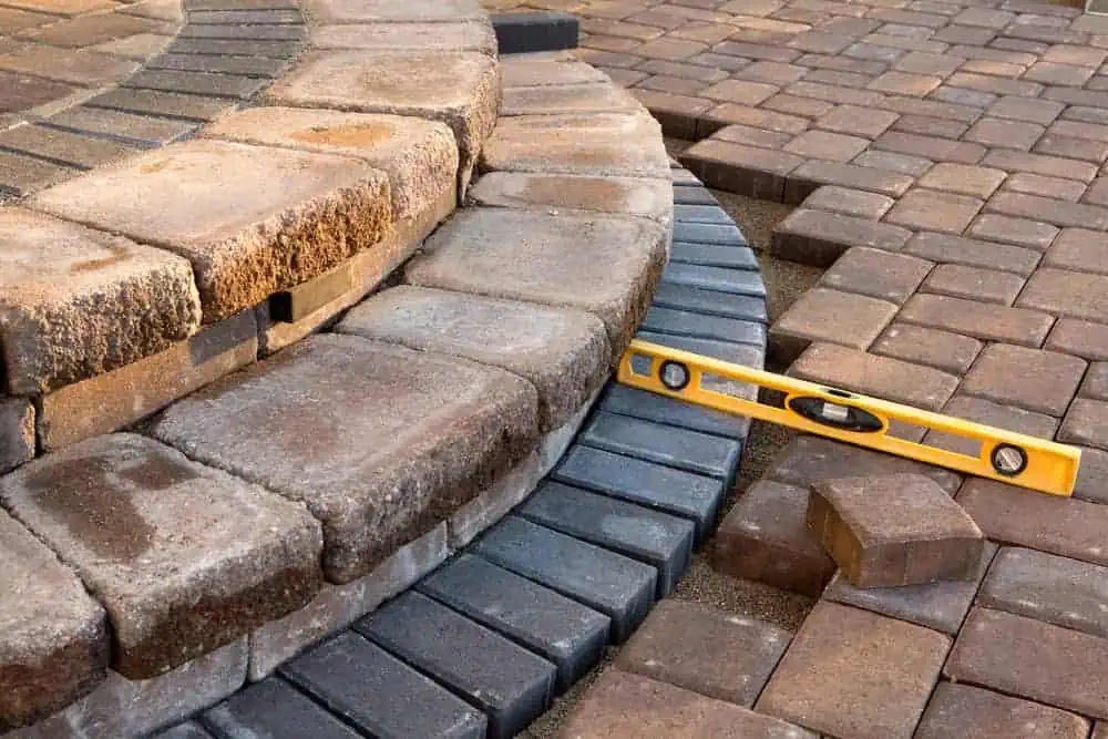 A close-up of a partially constructed brick patio featuring curved stone steps and rectangular pavers, highlighting expert stone masonry in Boston. An orange carpenter's level rests on the pavers, indicating ongoing leveling and alignment of the stones. Sand is visible between the pavers.
