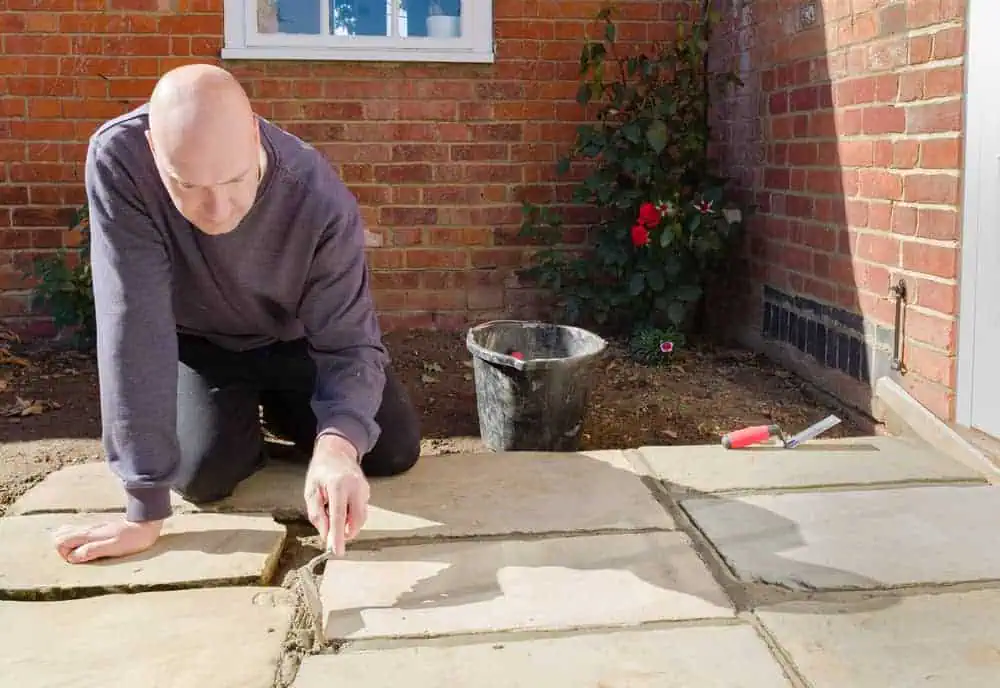 A person with a bald head in a grey sweatshirt is crouched down, applying or fixing grout between large stone slabs on a patio. A red brick house wall with a window and a rose bush is in the background, and a black bucket and some tools are beside them—an example of detailed Stone Masonry Boston work.