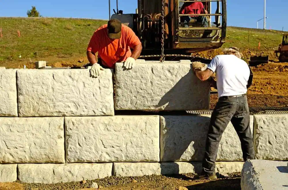 Two construction workers position large concrete blocks, building a retaining wall at a construction site. One worker in an orange shirt and black cap looks down, while the other in a white shirt adjusts the block from below. A digger and other equipment are in the background, showcasing Stone Masonry Boston expertise.