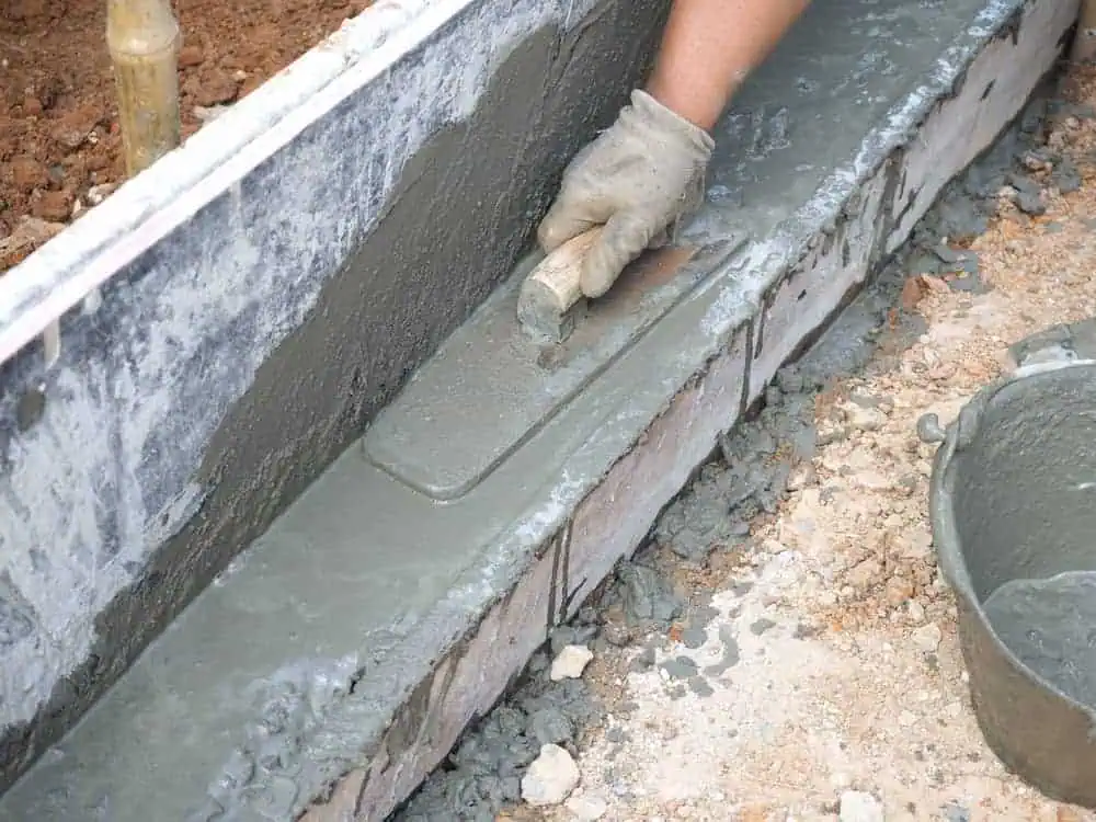 A person wearing a glove uses a trowel to smooth freshly applied cement on a construction site. The cement, crucial for Foundation Repair Boston projects, is being spread over a layer of bricks, with a bucket of additional cement visible on the side. The surface around the area is rough and unfinished.