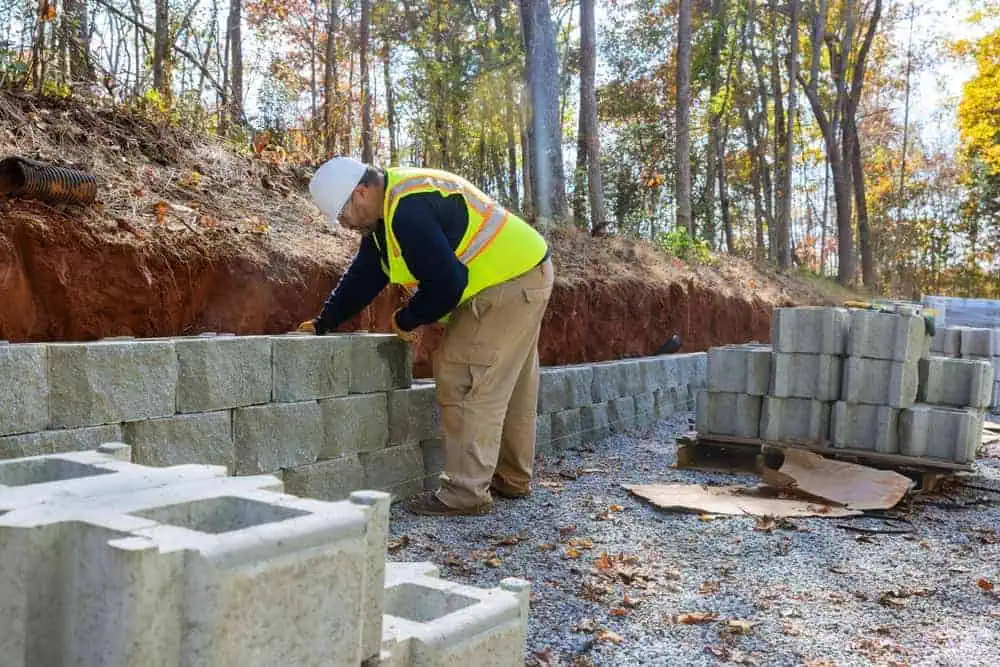 A construction worker wearing a hard hat and safety vest is building a stone wall outdoors, laying large concrete blocks on a gravel surface against an earth bank. Amidst the surrounding trees with autumn foliage, his expertise in Paver Installations Boston ensures the structure's stability.