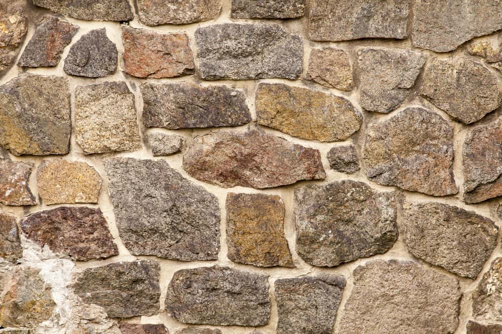 A close-up view of an old stone wall made of irregularly shaped rocks of various sizes and colors, including shades of brown, grey, and beige. The stones are fitted together with mortar, showcasing expert Stone Masonry Boston craftsmanship and giving the wall a rustic, textured appearance.