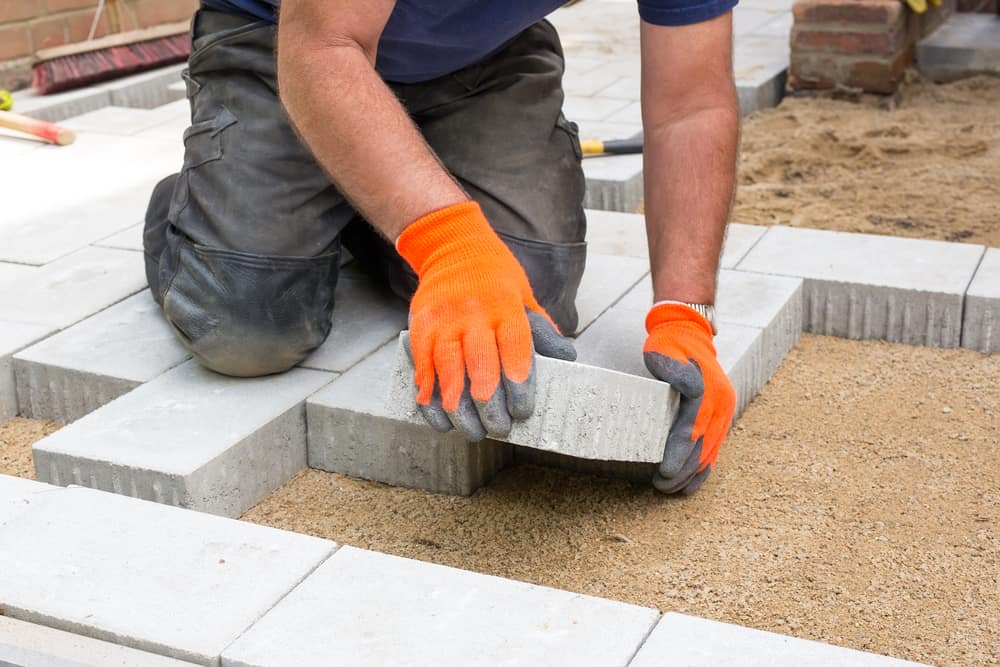 A person wearing bright orange gloves is placing a concrete paver onto a sandy surface while constructing a pavement or walkway. The Stone Masonry Boston expert is kneeling and surrounded by other pavers, showcasing their meticulous work in Paver Installations Boston.