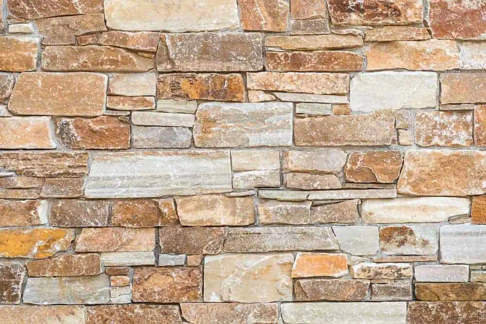 A close-up image of a stone wall made from irregularly shaped and variously colored stones, including shades of brown, tan, and gray. The stones are arranged in a rough, mortarless pattern, creating a textured and rustic appearance perfect for Paver Installations Boston.