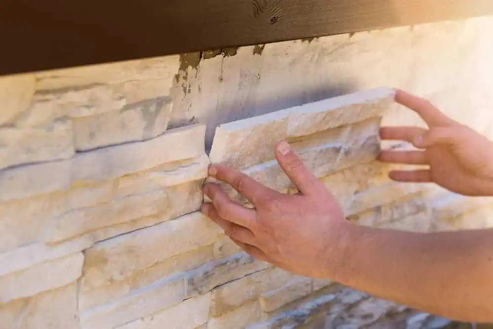 Close-up of hands installing beige stone veneer panels onto an outside wall, with one panel being pressed into place. The partially finished wall showcases the natural stone texture, with adhesive visible behind the panel, similar to techniques used in paver installations Boston professionals recommend.