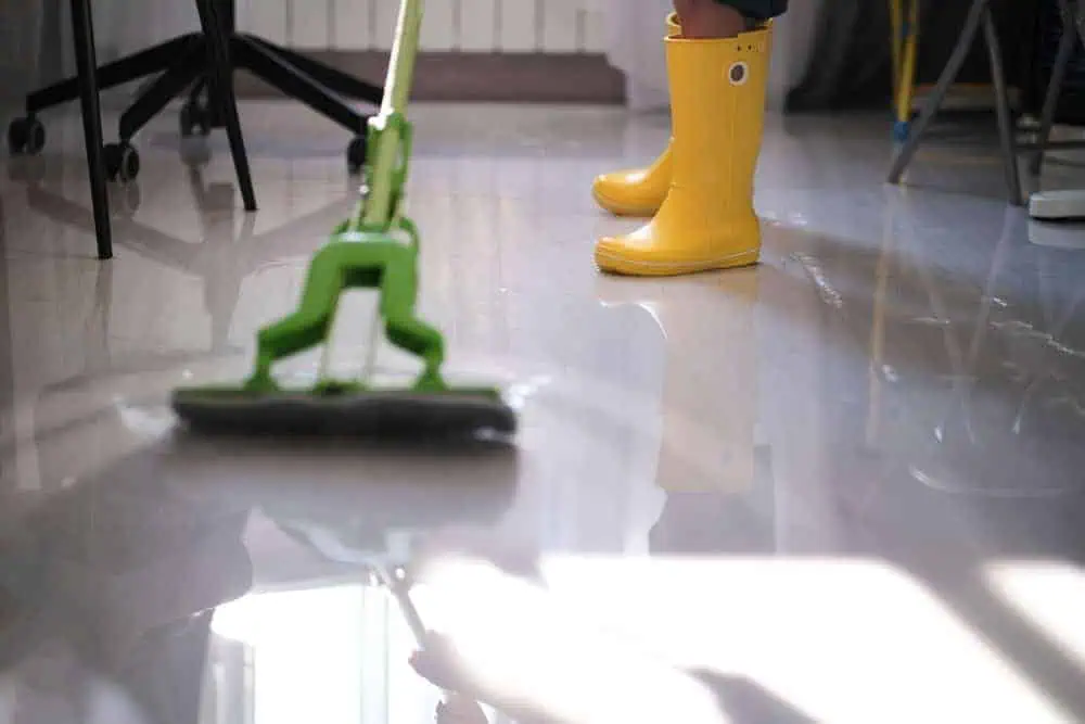 A person mopping a shiny floor with a green mop handle and wearing yellow rain boots. The reflection of the mop and boots gleams on the wet floor, alongside glimpses of furniture legs in the background, reminiscent of the precision you'd expect from a top-tier paving contractor in Middlesex County.