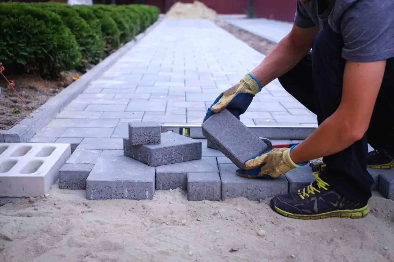 A worker wearing gloves and sneakers places rectangular concrete pavers onto a partially completed pathway. Sand and additional pavers are nearby, and green bushes line one side of the path. The image highlights precise outdoor paving work, typical of a Suffolk County paving contractor.