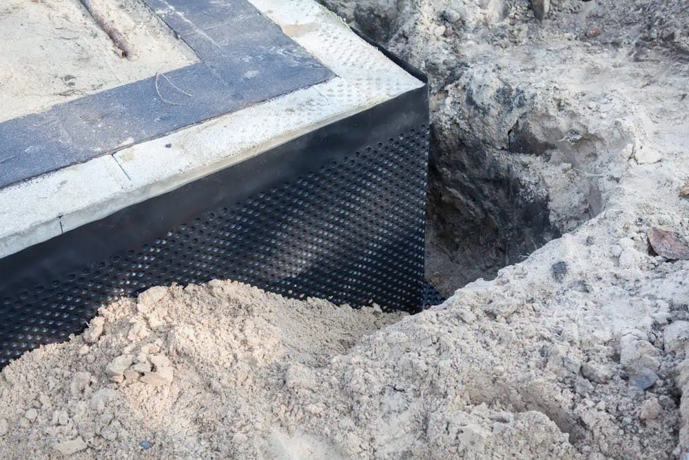 A section of a building foundation is shown with newly installed waterproofing. The foundation wall has a black, textured waterproof membrane attached, and there's excavated dirt around the area. The scene indicates ongoing construction or repair work by a masonry contractor in Suffolk County.