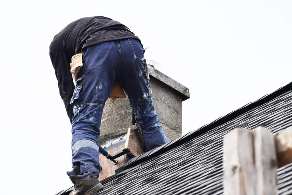 A worker in blue pants and a black shirt is installing or repairing roofing shingles on a sloped roof. Partially kneeling and bending over near the chimney, likely for Chimney Repair Suffolk County, tools and a tool belt are visible around their waist.