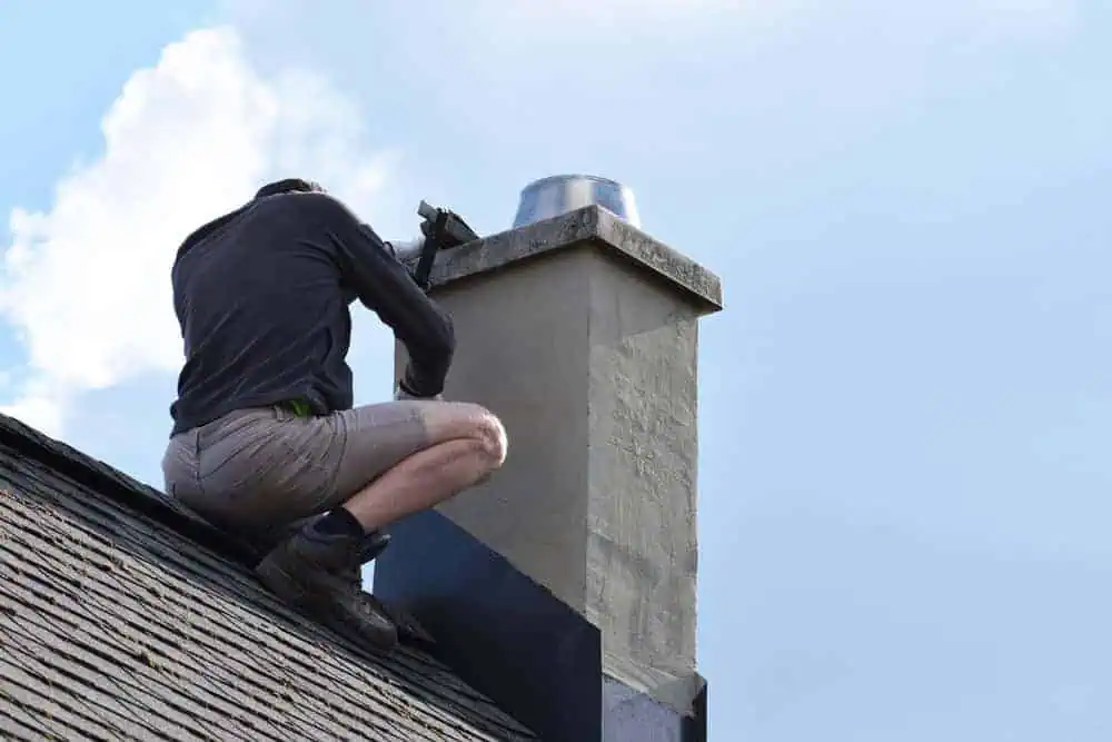 A person wearing a black jacket and beige shorts is kneeling on a rooftop, performing chimney repair in Middlesex County. The sky is clear with a few clouds visible in the background.
