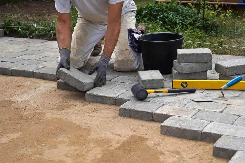 A person wearing gloves and a white shirt is installing rectangular pavers on a sand base in an outdoor setting, much like what you'd expect from a professional paving contractor in Suffolk County. Nearby are a bucket, rubber mallet, spirit level, and trowel. The area is surrounded by greenery.