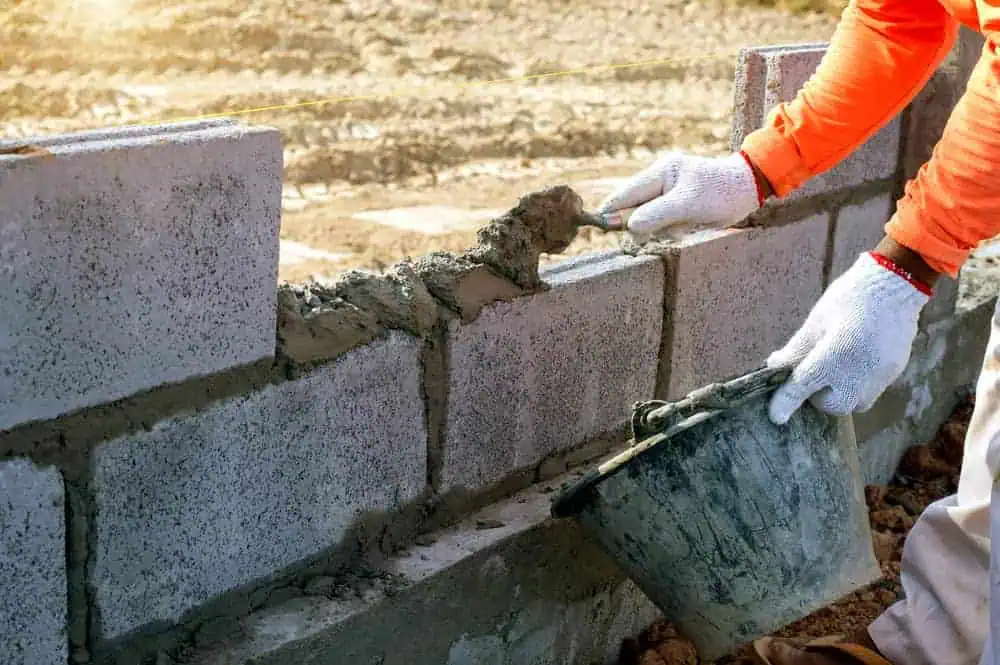 A person wearing a bright orange long-sleeve shirt and white gloves is applying mortar with a trowel to a concrete block wall they're constructing. They hold a bucket of mortar in one hand, and the wall is only a few blocks high. The background shows muddy ground typical for any Masonry Contractor Suffolk County project.