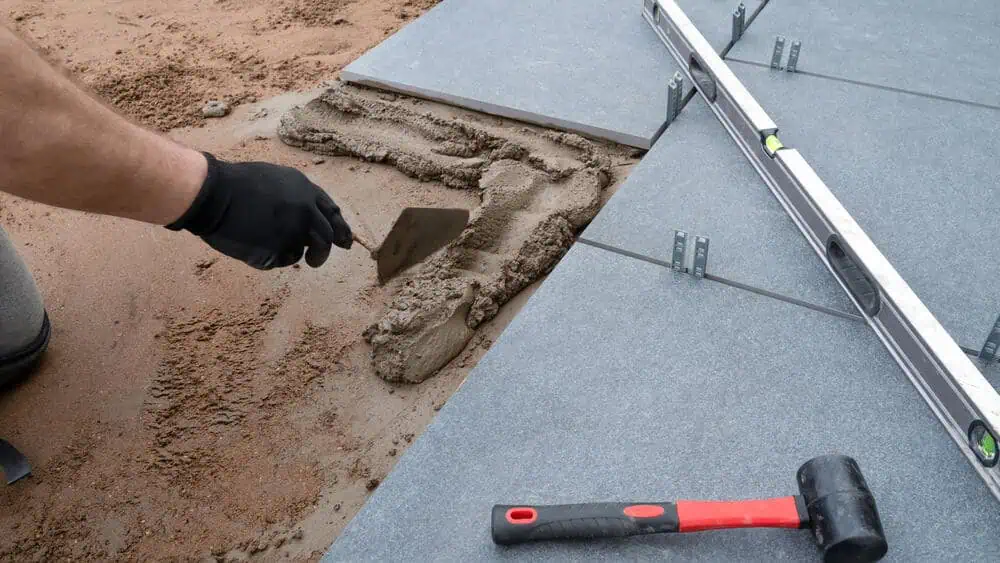A person wearing black gloves uses a trowel to spread cement on a sandy surface next to large, gray tiles. A rubber mallet and a leveling tool are placed nearby. The scene suggests the detailed work of a masonry contractor in Middlesex County specializing in outdoor tiles or paving stones.