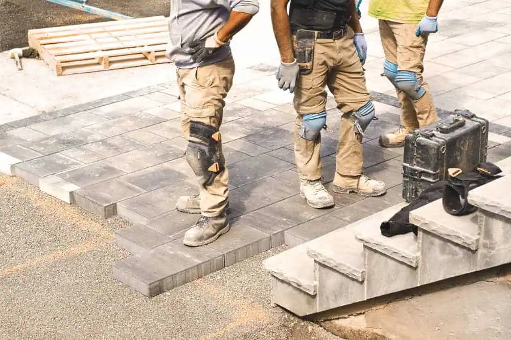 Three construction workers in work gear, including pants with kneepads, safety vests, and gloves, stand on newly laid concrete pavers near a stack of concrete blocks and tools. As experienced paving contractors in Middlesex County, they appear to be discussing their work on the pavement.