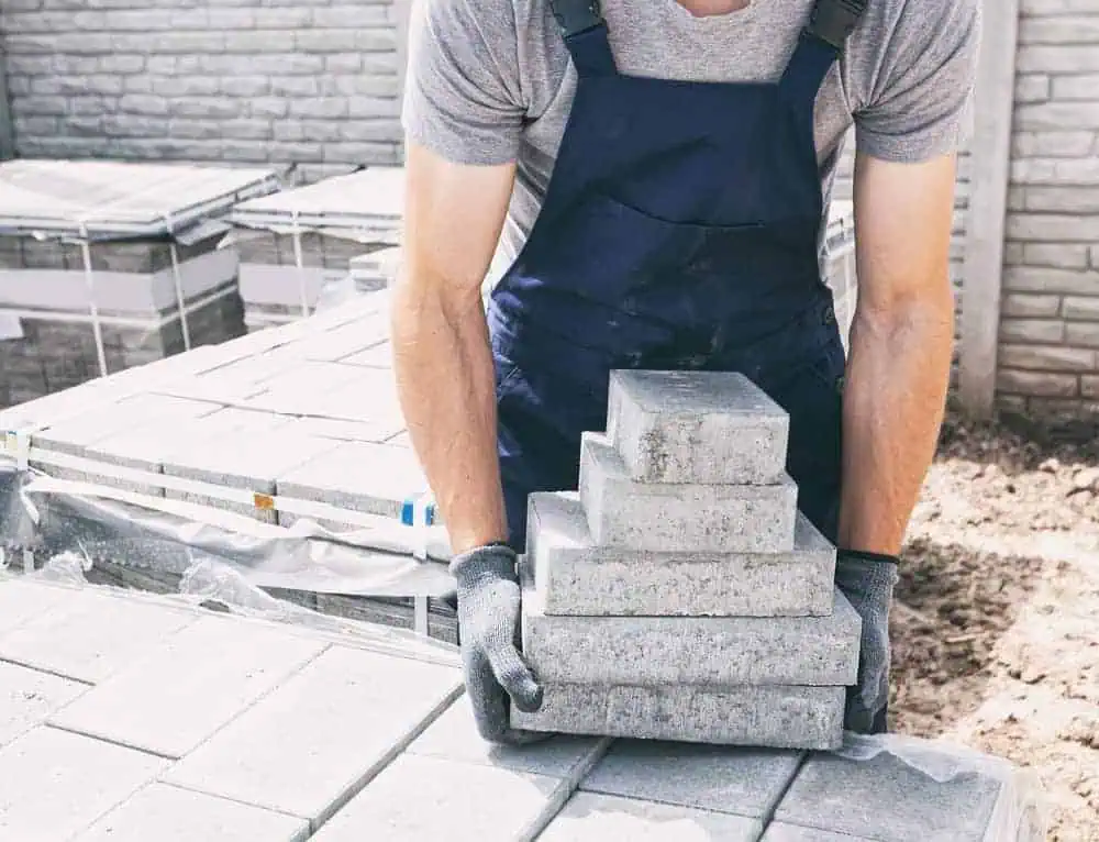 A worker wearing a grey t-shirt, blue overalls, and gloves is lifting a series of stacked concrete pavers. The background shows more pavers and a brick wall, typical of tasks completed by masonry contractors in Suffolk County. The worker's face is not visible.