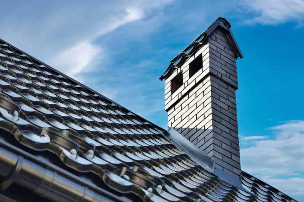 A close-up view of a house roof with shiny, wavy metal tiles and a brick chimney under a blue sky with a few clouds. The chimney has two openings at the top, highlighting the precision work typical of Chimney Repair Middlesex County.