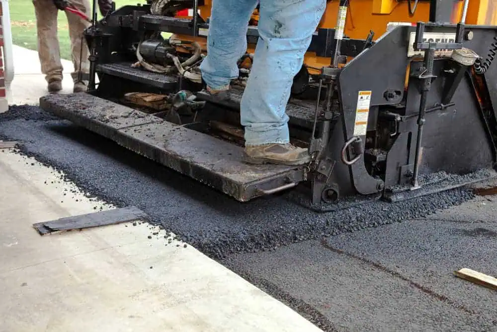 A close-up of a construction worker's legs and feet operating a heavy road paving machine. The worker is wearing blue jeans and boots. Fresh asphalt is being laid on the ground, and the machinery is applying it smoothly. Other workers are visible in the background, highlighting a Paving Contractor Suffolk County job.