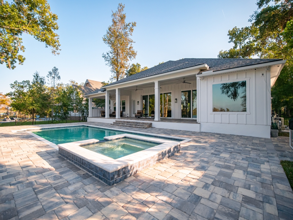 A modern white house with a large patio area and an outdoor swimming pool. The pool has a rectangular shape with a smaller, raised hot tub at one end. The yard is paved with light-colored bricks, and trees provide shade in the background.