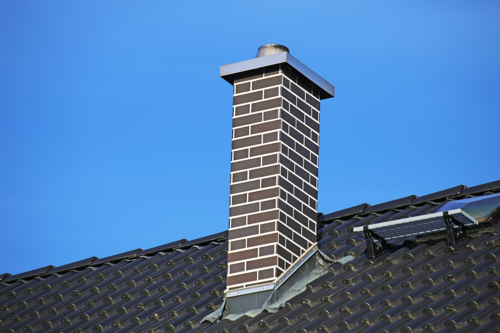 A brick chimney with a metal cap rises from a dark tiled roof against a clear blue sky. The roof also features a small solar panel.