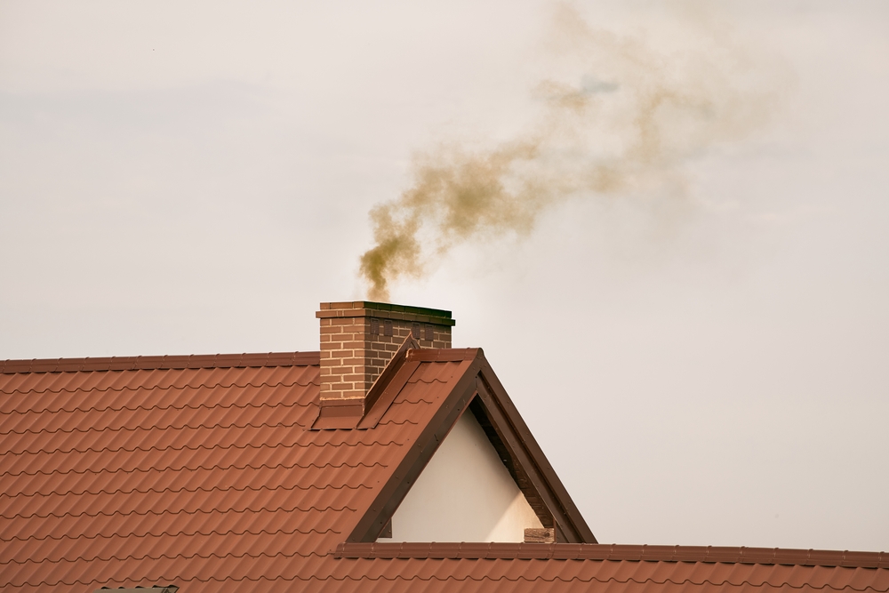 A sloped red-tiled roof with a brick chimney emits a plume of gray smoke against an overcast sky.