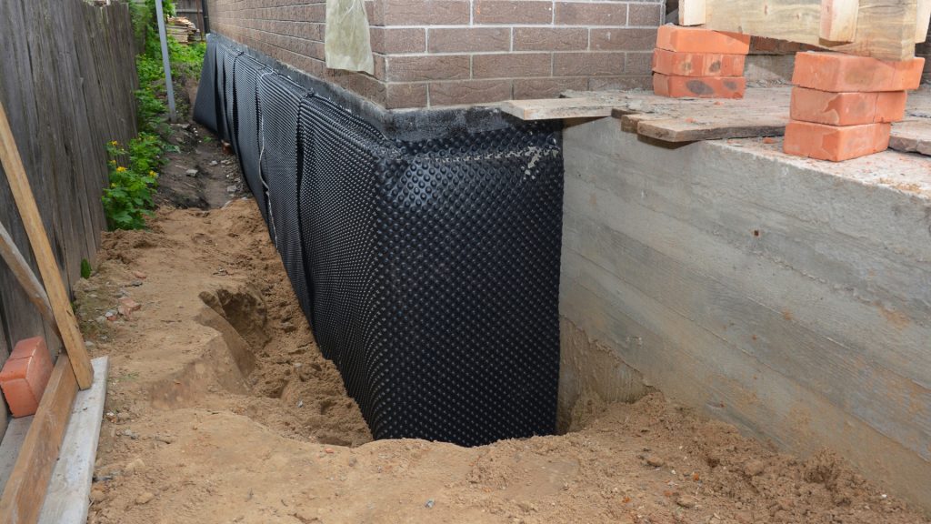 A close-up of a building foundation shows a layer of black waterproofing membrane installed outside the wall, with exposed soil and construction materials like bricks nearby. The area appears to be a construction site for foundation waterproofing.