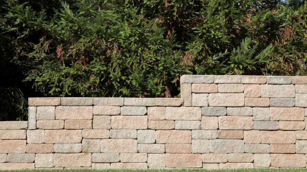 A stone retaining wall made of rectangular blocks stands in front of lush green foliage on a sunny day.