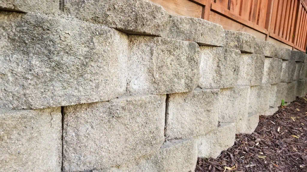 Stone retaining wall with large, rectangular blocks stacked in a staggered pattern. The wall borders a wooden fence above, with mulch covering the ground at the base.
