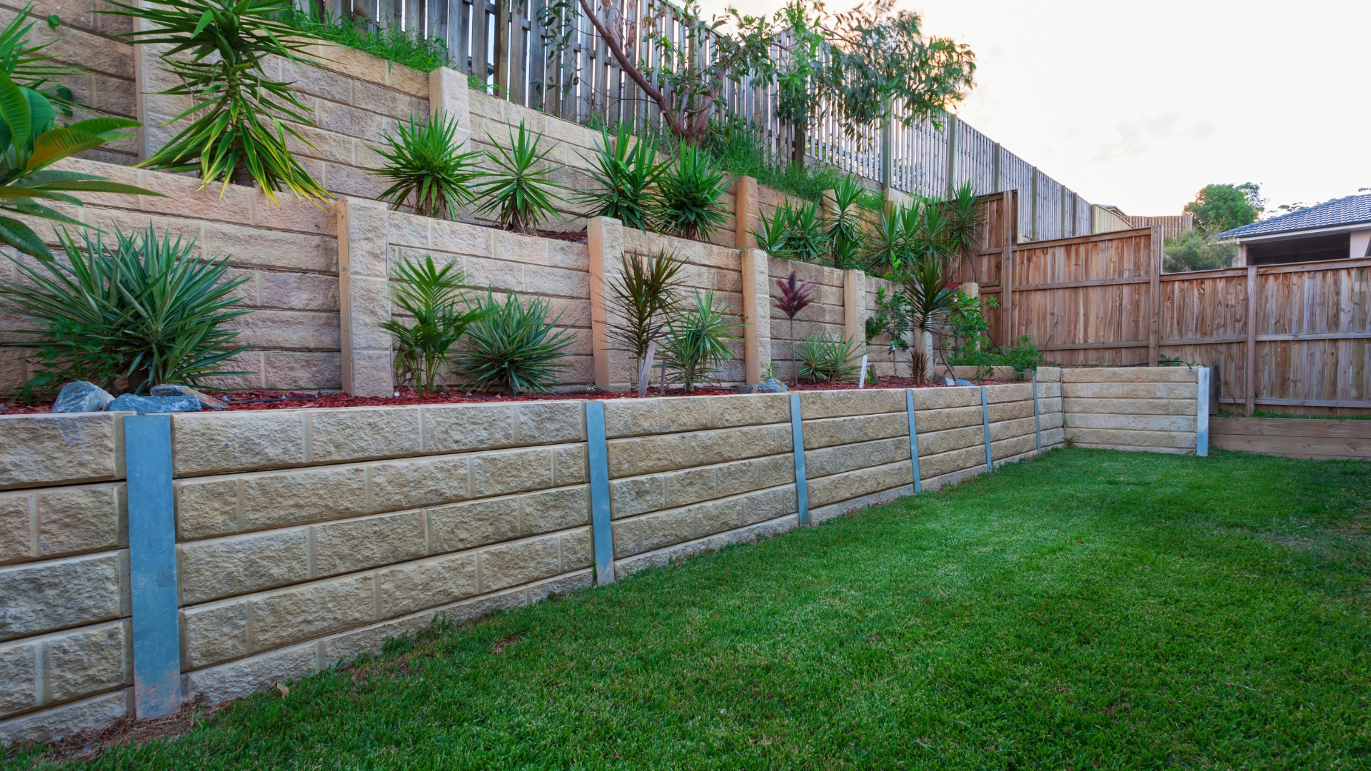 A landscaped backyard featuring tiered stone retaining walls planted with green and red tropical plants. The grass lawn is well-maintained, and a wooden fence runs along the top of the slope. The image conveys a peaceful, organized, and aesthetically pleasing outdoor space.