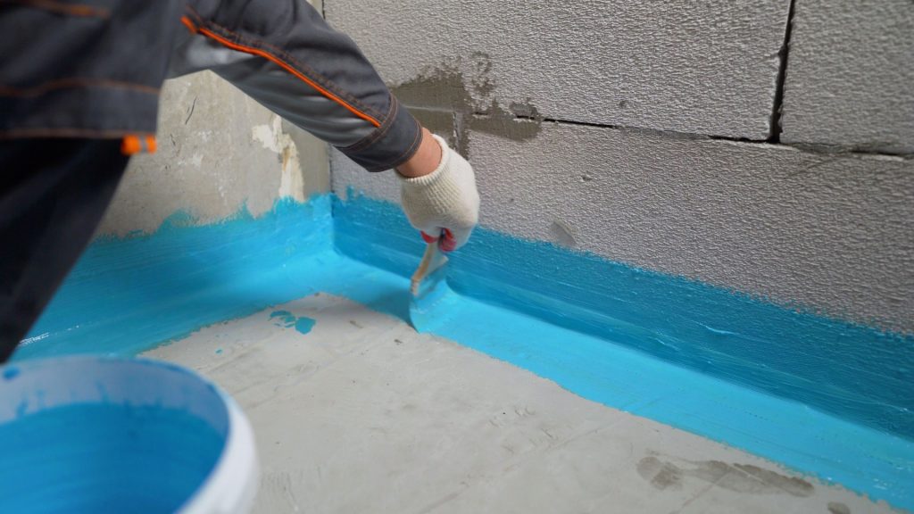 A person wearing gloves applies blue waterproofing paint with a brush to the corner of a room with gray concrete walls. A bucket with more paint is seen beside them.