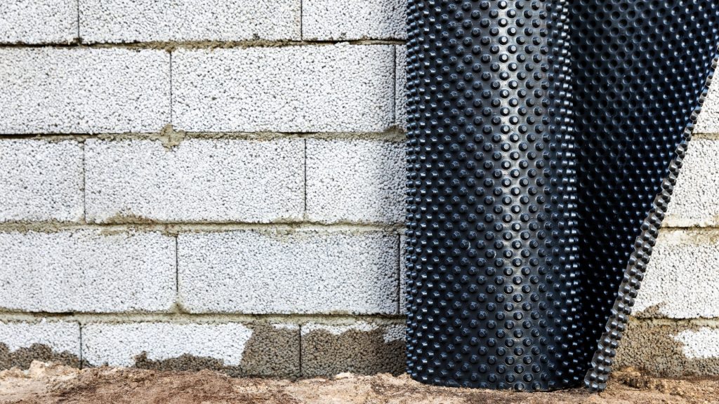 A close-up of a brick wall with a section of black dimpled drainage membrane partially covering it. The ground is bare, and the membrane is used for waterproofing and protecting the foundation.