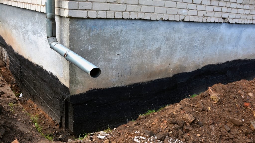 A building corner with a visible rain gutter pipe is shown. The foundation is coated with a black waterproofing layer above exposed earth in front of the façade. The building has a white brick wall above a concrete base.