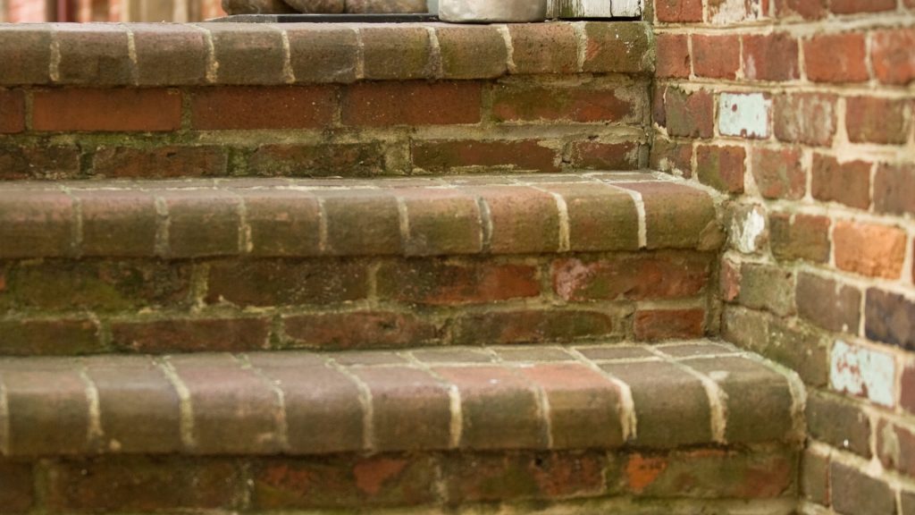 Close-up view of an old brick staircase with visible moss and weathering, showcasing a worn and rustic appearance. The bricks are laid in a stretcher bond pattern, and the steps lead upwards, partially framed by a matching brick wall.