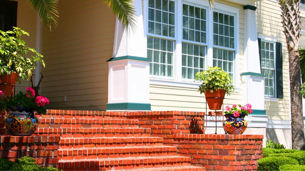 A sunlit, yellow house with large windows and white trim. Brick steps lead to the entrance, adorned with colorful potted plants and flowers. Palm tree leaves frame the scene, creating a vibrant, inviting atmosphere.