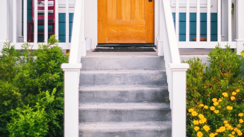 Concrete steps lead up to a wooden front door with white railings on each side. Green shrubs and bright yellow flowers border the steps, creating a welcoming entrance to the home.