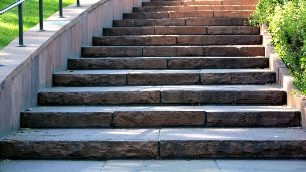 A stone staircase with weathered steps ascends flanked by simple metal railings on the left and greenery on the right, bathed in sunlight.