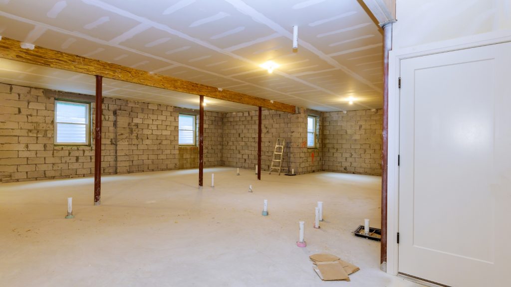 A spacious unfinished basement with concrete walls and floor, exposed beams, and ceiling lights. There are several PVC pipes protruding from the ground, two windows on the left wall, a metal ladder in the back, and a white door on the right.