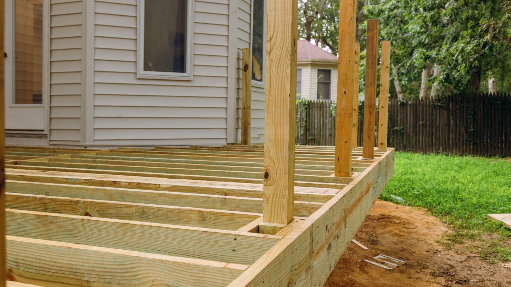 A wooden deck under construction extends from the back of a house. Several upright wooden posts are in place, with visible joists forming the deck's framework. The area is surrounded by grass and a privacy fence.