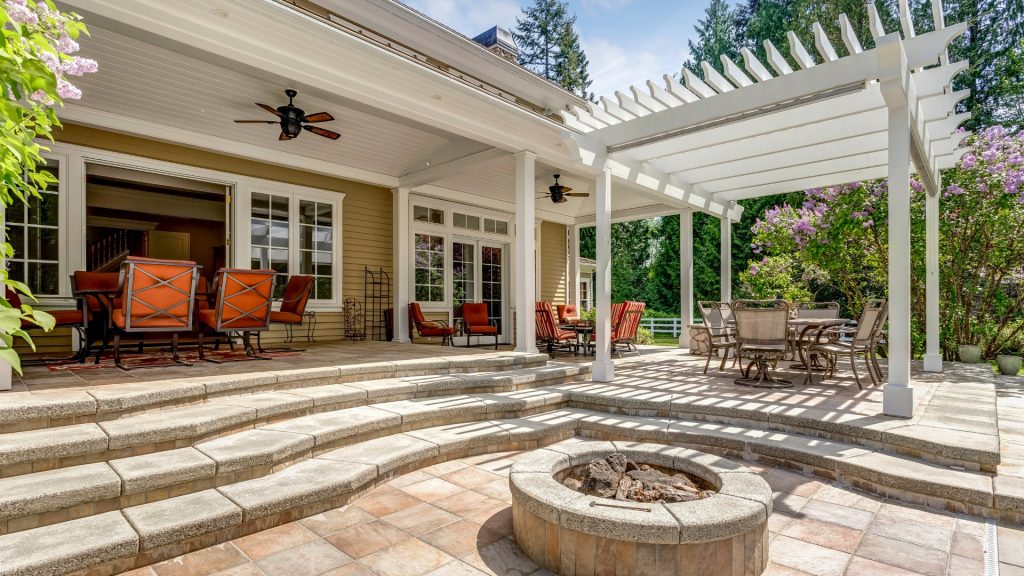 A spacious outdoor patio with a fire pit, multiple seating areas, and a pergola casting shadows on the tiles. There are red cushioned chairs around dining tables and steps leading to a house with glass doors. Lush greenery surrounds the area.