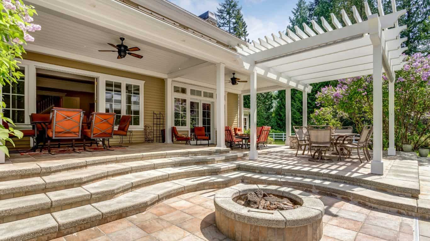 A spacious outdoor patio featuring a fire pit and multiple seating areas. A pergola casts shadows on the tiled floor, and red-cushioned chairs surround dining tables. Steps lead to a house with glass doors, and lush greenery surrounds the patio, creating an inviting and well-designed outdoor living space.
