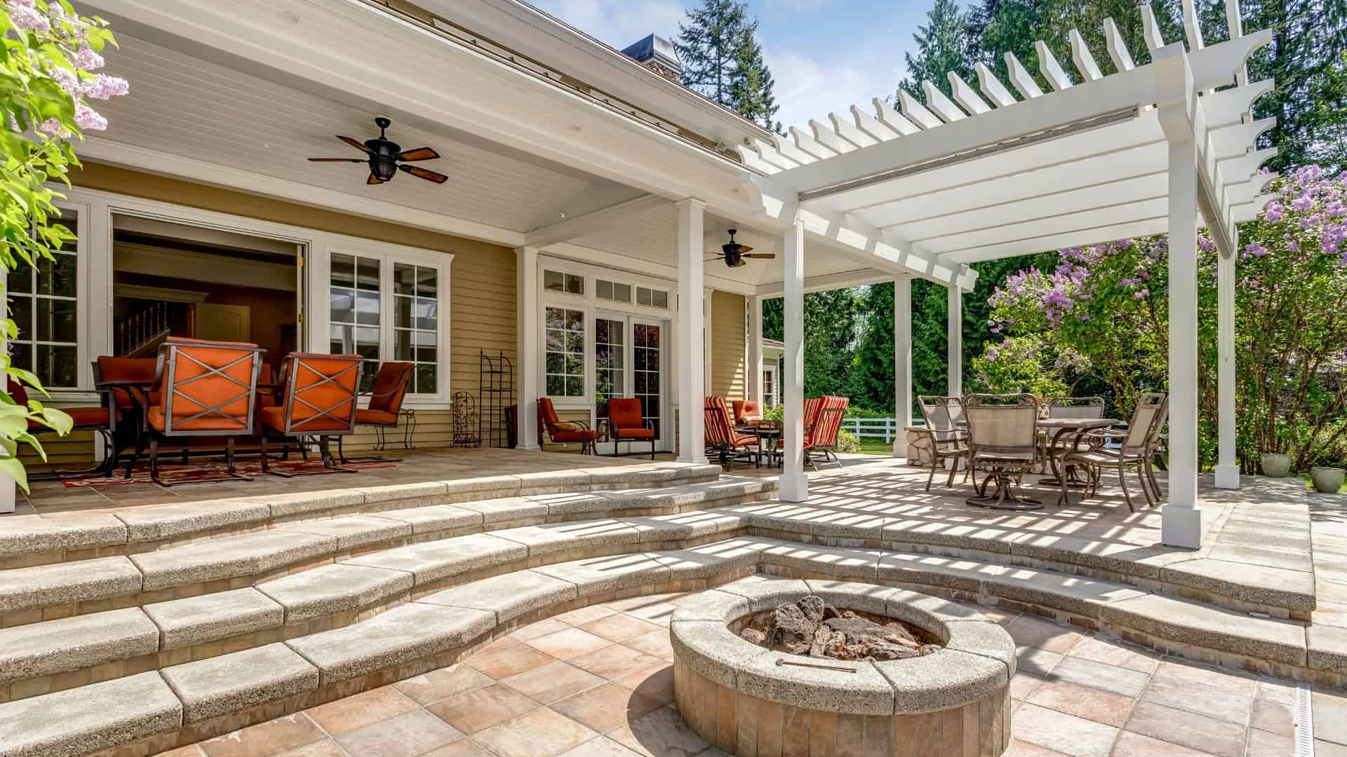 A spacious outdoor patio with a fire pit, multiple seating areas, and a pergola casting shadows on the tiles. There are red cushioned chairs around dining tables and steps leading to a house with glass doors. Lush greenery surrounds the area.
