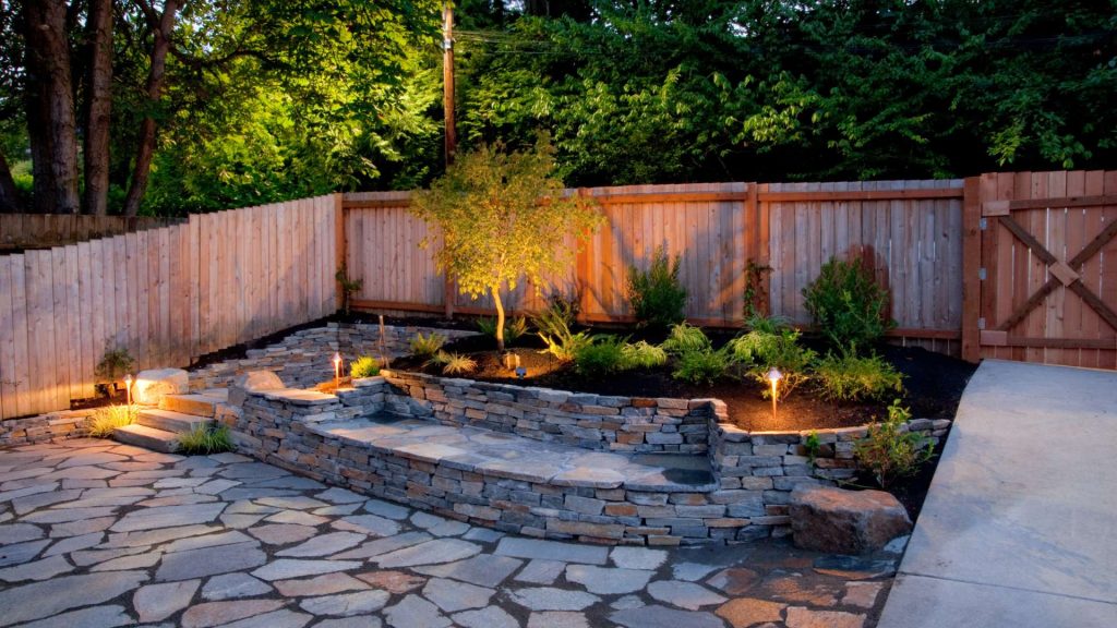 A serene backyard features a stone patio with warm, ambient lighting. A stone retaining wall encloses a small garden with shrubs and a tree. A wooden fence borders the yard under a canopy of green trees.