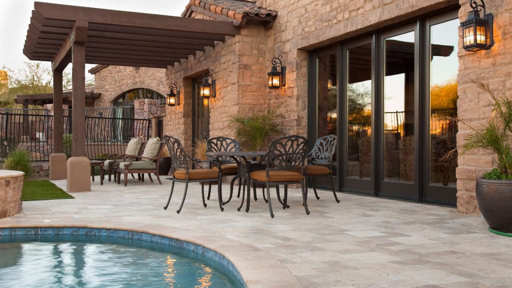 A stone patio with a round table and four chairs next to a pool. The wall has large glass doors and lit wall lanterns. A pergola extends over a seating area with cushioned chairs, creating a cozy outdoor space.